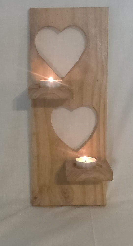 A rustic wooden wall-mounted candle holder with two heart-shaped cutouts and two small shelf levels holding lit tea light candles, shown against a plain light fabric background.